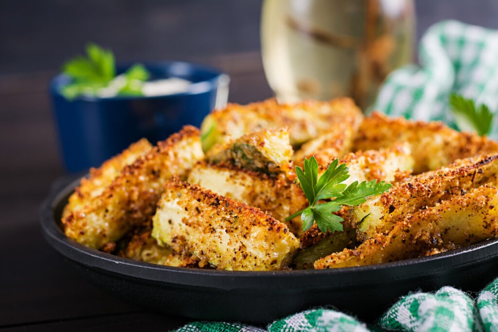 Knusprige Zucchini-Pommes mit Parmesan-Kruste auf einem BackblechBeschreibung: Goldbraune, knusprige Zucchini-Pommes mit würziger Parmesan-Mandel-Kruste, serviert auf einem rustikalen Backblech mit einem cremigen Dip daneben.